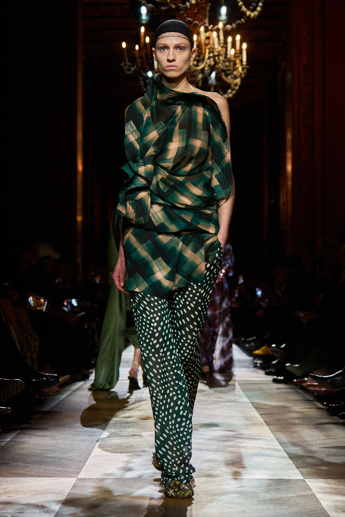 Model walks a runway in a green plaid draped top and polka-dot pants, with chandeliers lit behind her in a fashion show setting.