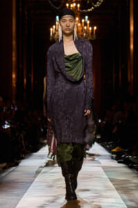 Model on a runway wearing a dark purple patterned coat over a draped green top, accessorized with long white earrings and a black headscarf; chandeliers glow in the background.