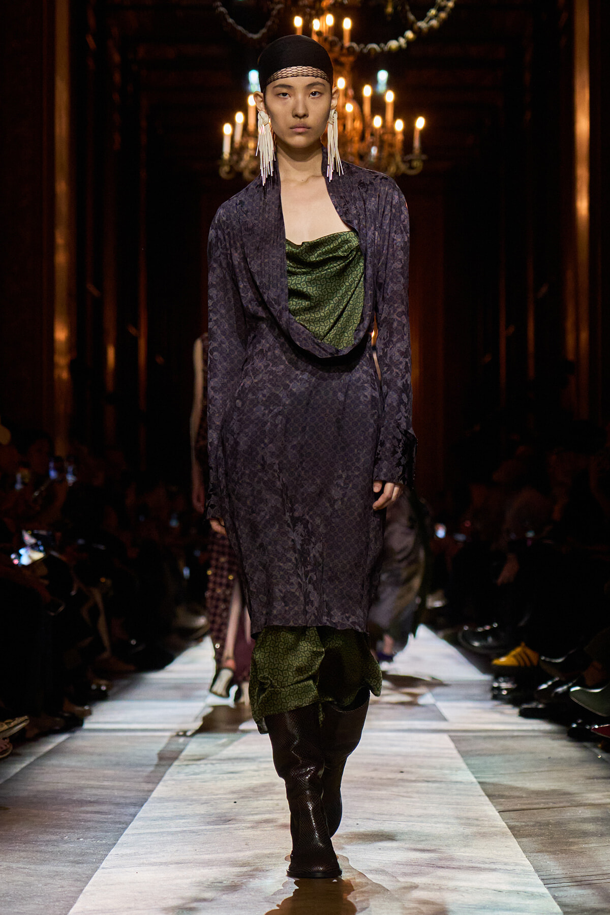 Model on a runway wearing a dark purple patterned coat over a draped green top, accessorized with long white earrings and a black headscarf; chandeliers glow in the background.