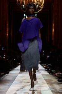 Model walks a runway in a cobalt purple draped top and a gray shaggy midi skirt, white shoes, chandeliers in the background.