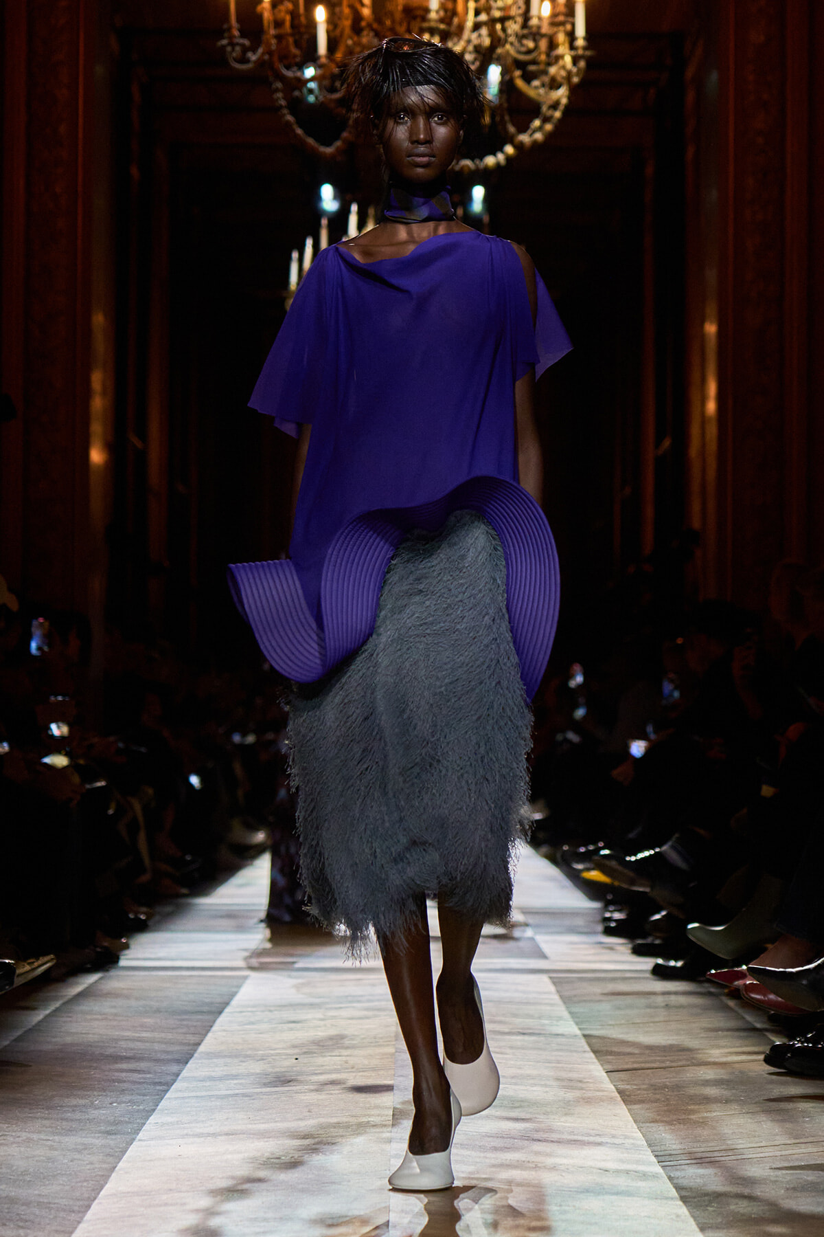 Model walks a runway in a cobalt purple draped top and a gray shaggy midi skirt, white shoes, chandeliers in the background.