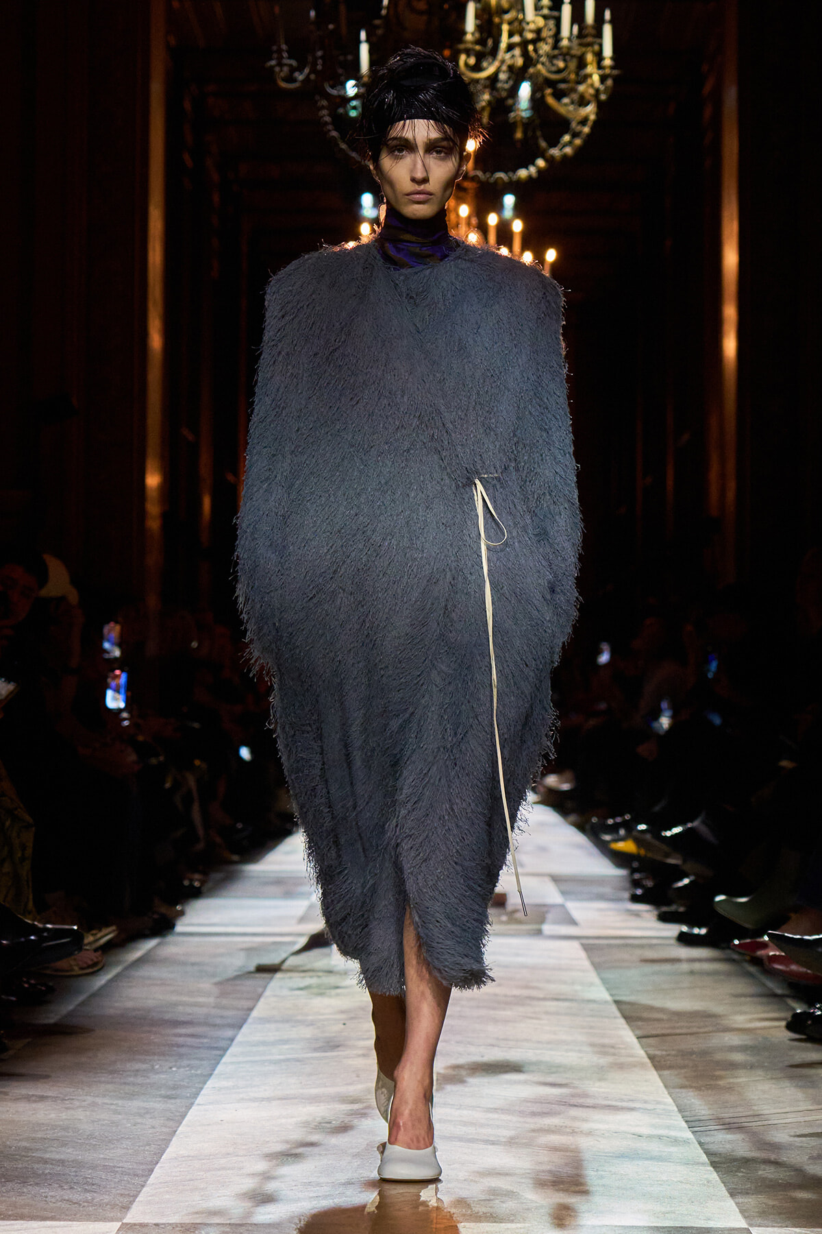 Model on a runway wearing a loose blue shaggy coat with a white cord belt and white heels, chandeliers glowing behind.