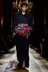 Model on a runway wearing a dark oversized coat with a multicolored tassel belt grading across the waist area.