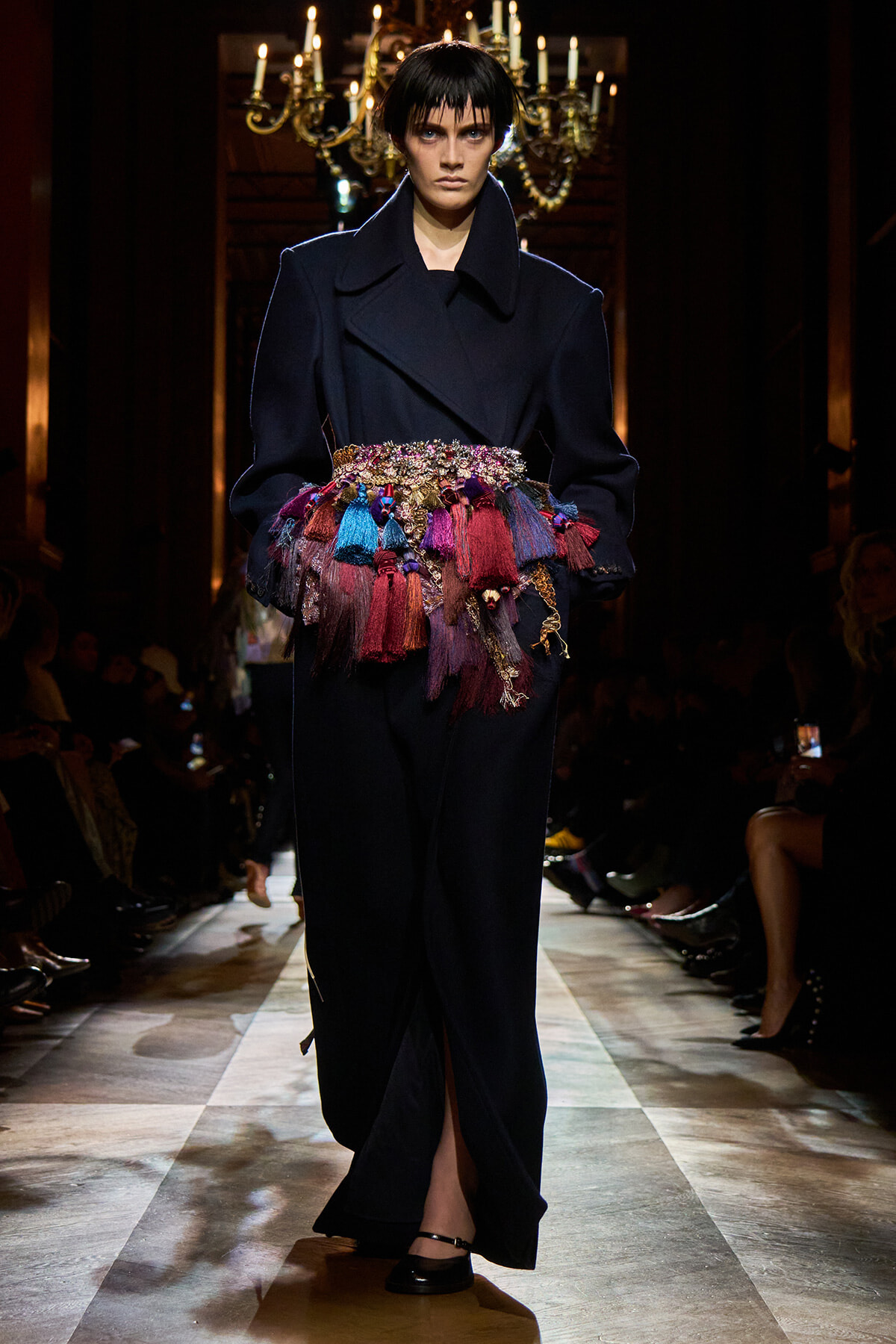 Model on a runway wearing a dark oversized coat with a multicolored tassel belt grading across the waist area.