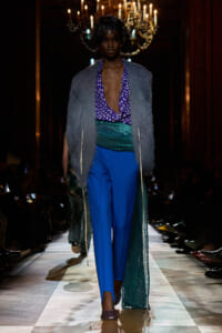Runway model wearing a purple polka‑dot deep V blouse, bright blue trousers, metallic green sash, and a gray fur vest on a lit catwalk.