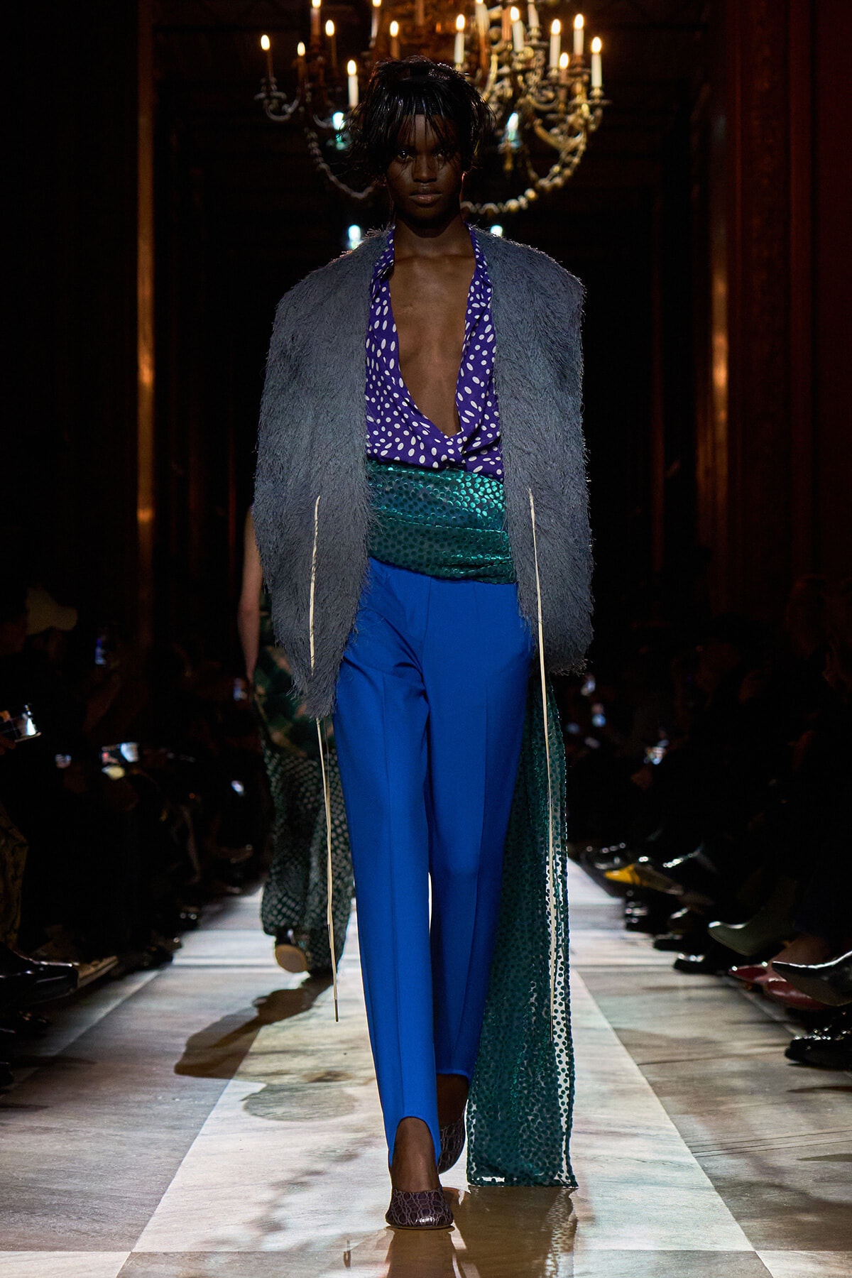 Runway model wearing a purple polka‑dot deep V blouse, bright blue trousers, metallic green sash, and a gray fur vest on a lit catwalk.