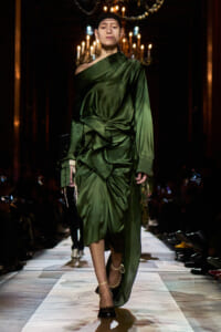 Model walks a fashion runway in a green satin draped dress with ruched detailing and an off-shoulder look, beige ankle-strap heels, chandeliers in the background.