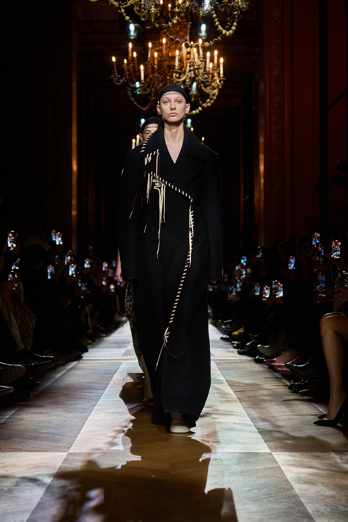 Model walks the runway in a long black coat with white rope trim, under ornate chandeliers in a dim, gilded hall.