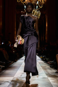 Model walks a runway in a dark purple satin one-shoulder gown, holding a colorful tassel clutch beneath chandeliers.