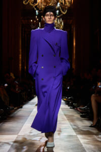 Model walks the runway in a cobalt purple double-breasted coat with strong shoulders and a high neck, hands in pockets.