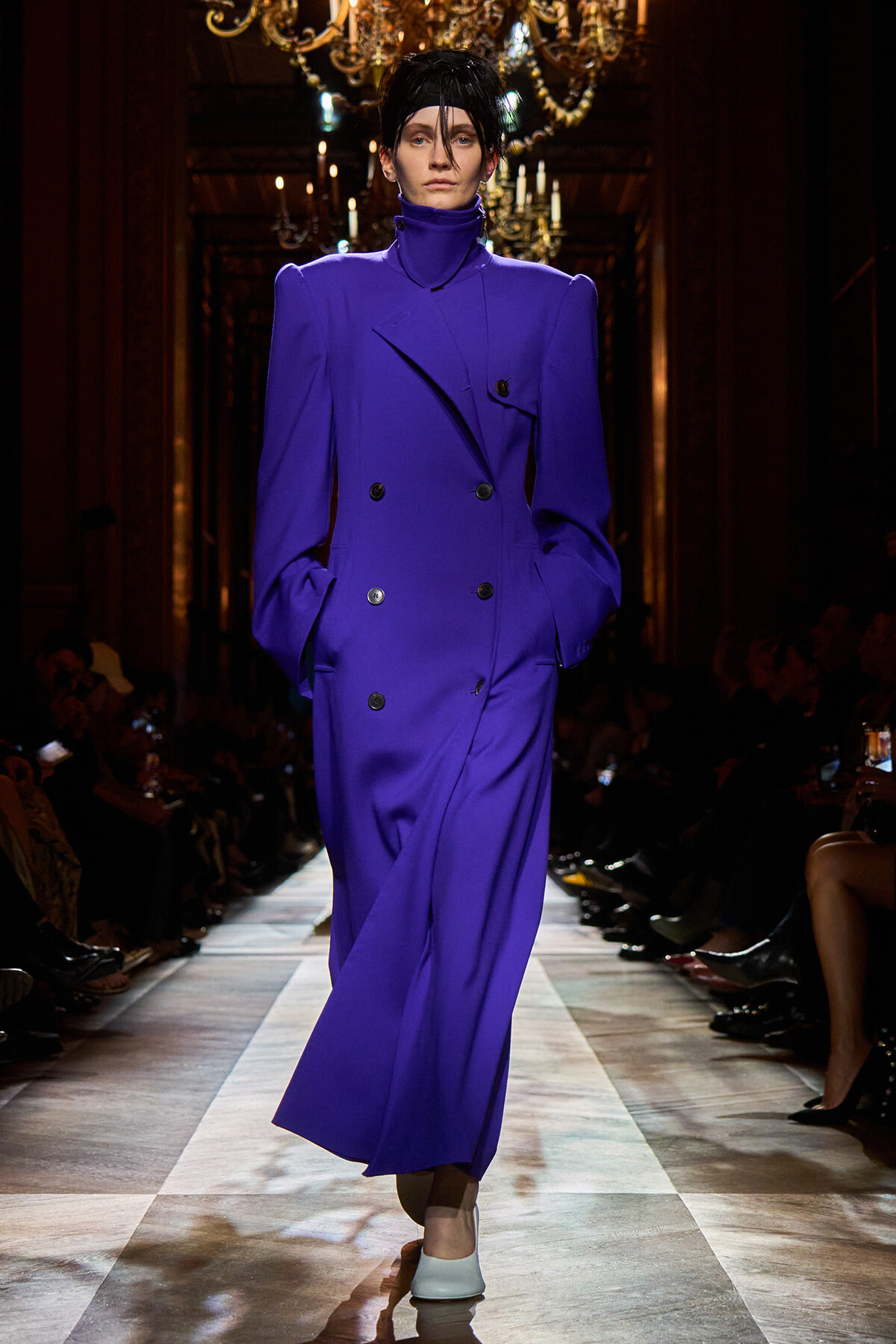 Model walks the runway in a cobalt purple double-breasted coat with strong shoulders and a high neck, hands in pockets.