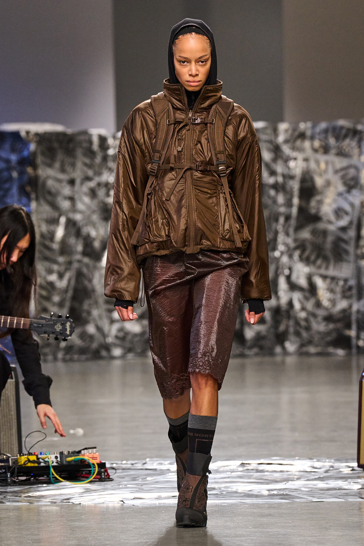 Model on a runway wearing a brown hooded windbreaker with a chest harness over matching brown shorts, paired with patterned boots and gray socks.
