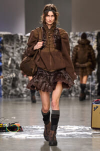 Model walking a runway in a brown, layered outfit with a lace skirt and headband, carrying a brown shoulder bag.