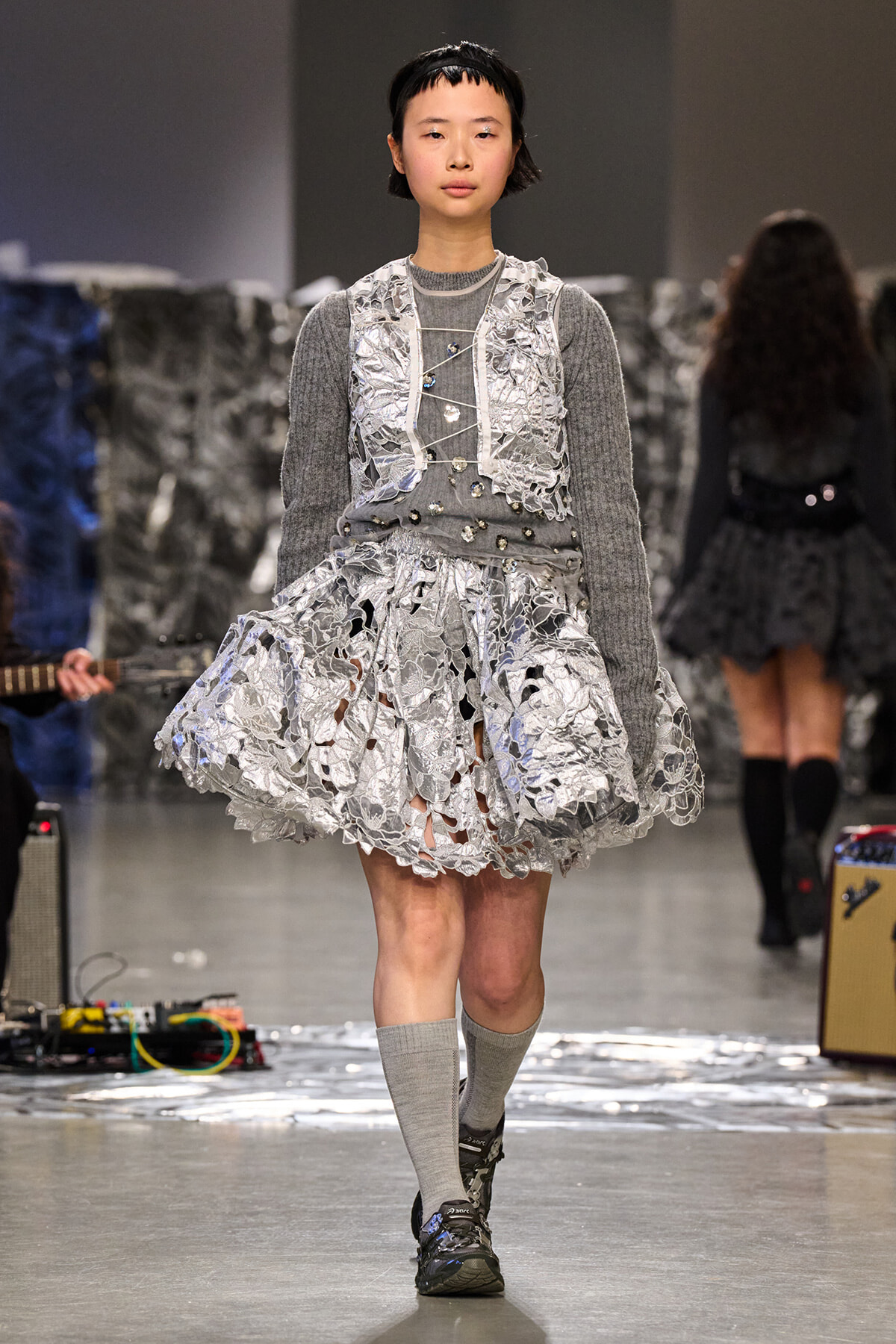 Female model on a runway wearing a gray knit top under a silver lace overlay dress with a voluminous skirt and gray knee-high socks, sneakers visible at feet.