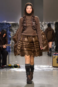 Model walking a runway in a brown ribbed knit sweater, oversized quilted puffer skirt with lace trim, and a matching brown backpack and headband.