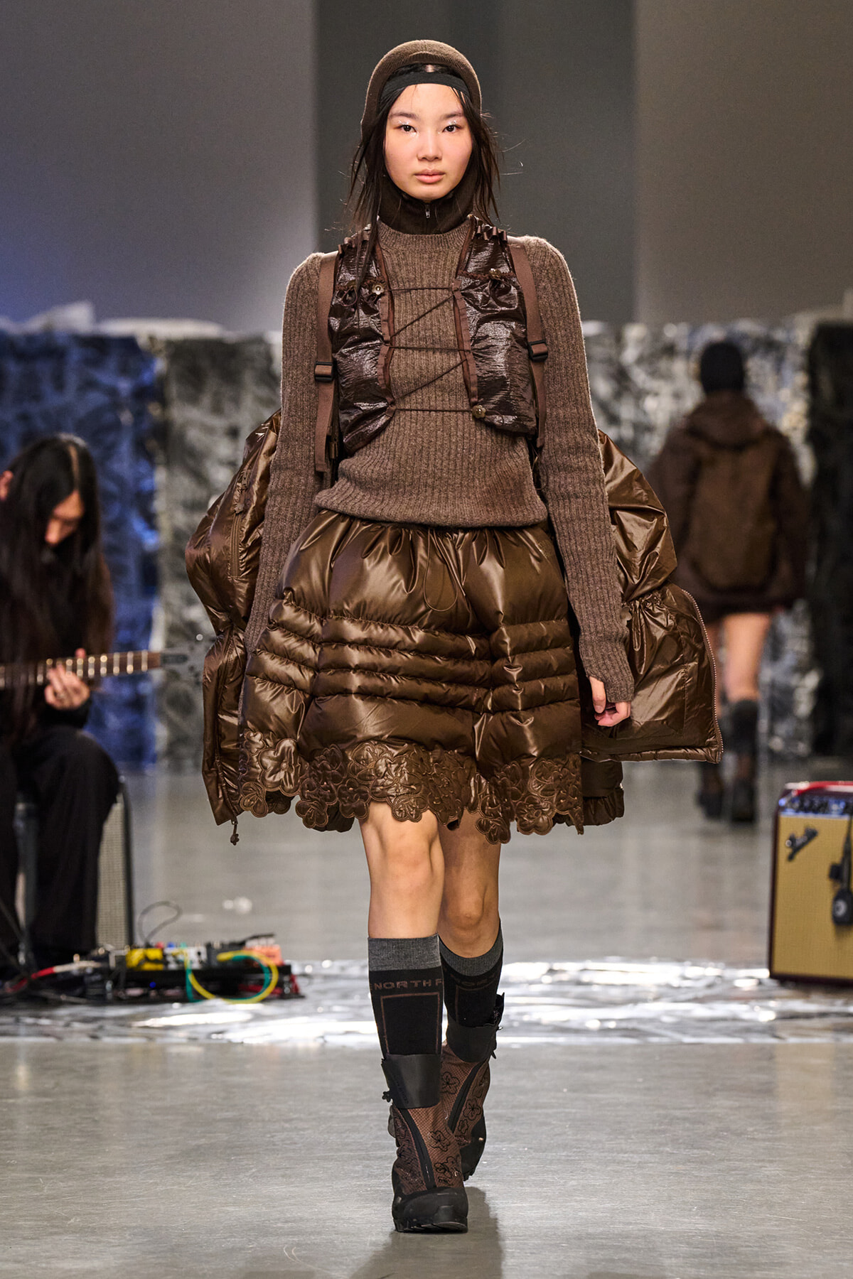 Model walking a runway in a brown ribbed knit sweater, oversized quilted puffer skirt with lace trim, and a matching brown backpack and headband.
