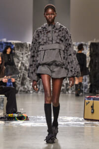 Model walking a fashion runway in a gray floral-embellished hooded pullover with a matching belt and short shorts, paired with black knee-high socks and floral shoes.
