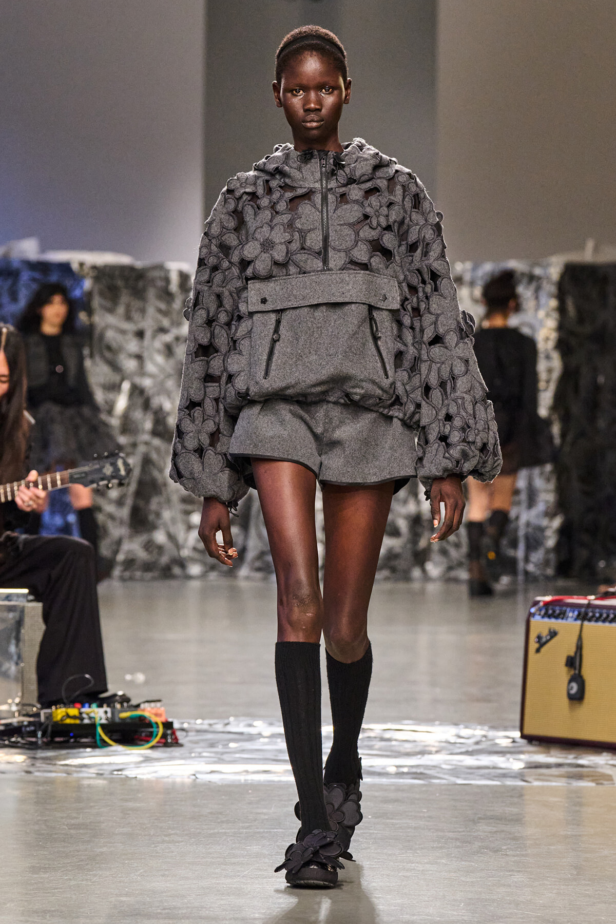 Model walking a fashion runway in a gray floral-embellished hooded pullover with a matching belt and short shorts, paired with black knee-high socks and floral shoes.
