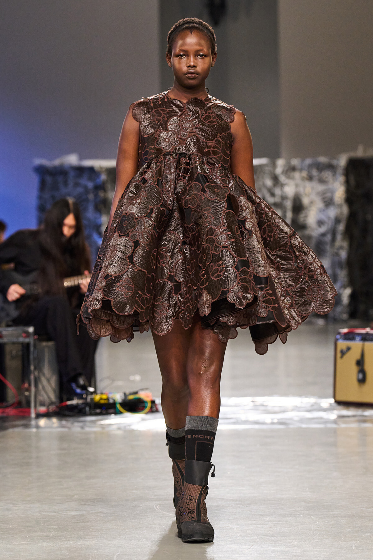 Female model walking runway in a brown lace floral-embellished dress with a voluminous skirt and ankle boots on a fashion show runway
