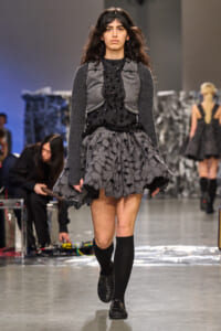 Female model walking a fashion show runway in a gray and black layered dress with a textured, leaf-patterned skirt and knee-high black socks under a headband.