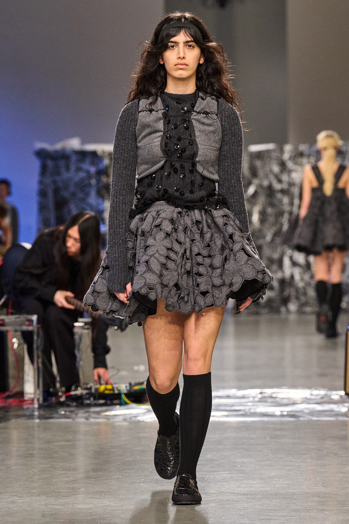 Female model walking a fashion show runway in a gray and black layered dress with a textured, leaf-patterned skirt and knee-high black socks under a headband.