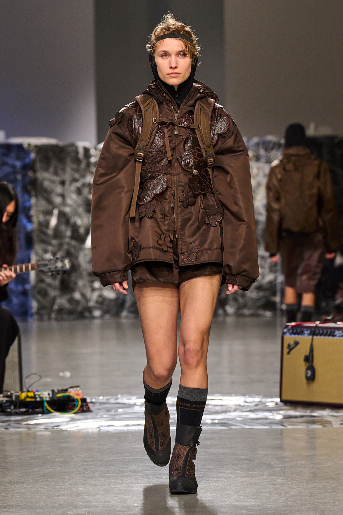 Model walks runway in oversized brown jacket with floral appliqués and backpack, brown boots on a fashion show stage.