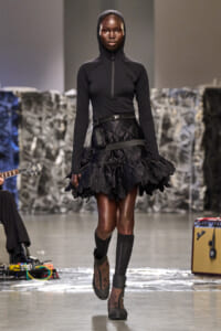Model walking runway in black hooded top with half-zip, a wide quilted belt, and a ruffled black skirt with floral lace edges; dark hosiery and ankle boots.