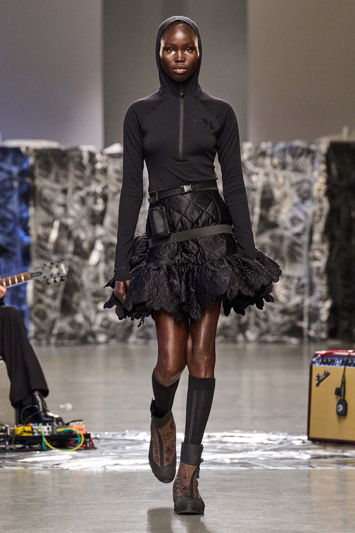 Model walking runway in black hooded top with half-zip, a wide quilted belt, and a ruffled black skirt with floral lace edges; dark hosiery and ankle boots.