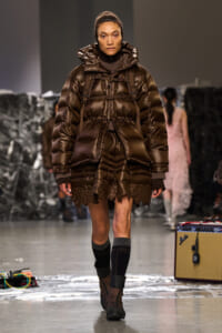 Model walks a runway in a glossy brown puffer coat with hood and lace-trim hem, paired with brown boots.