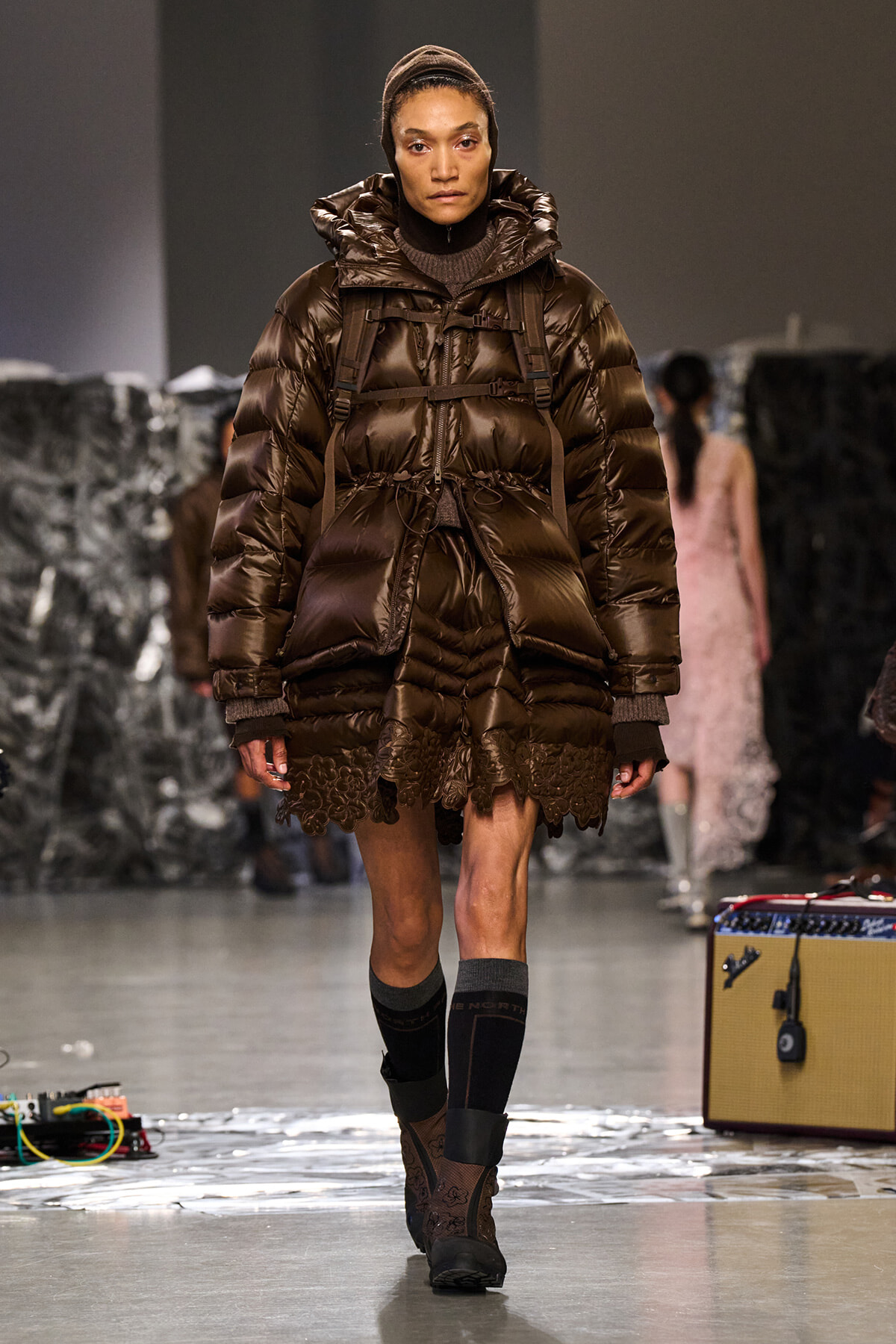 Model walks a runway in a glossy brown puffer coat with hood and lace-trim hem, paired with brown boots.