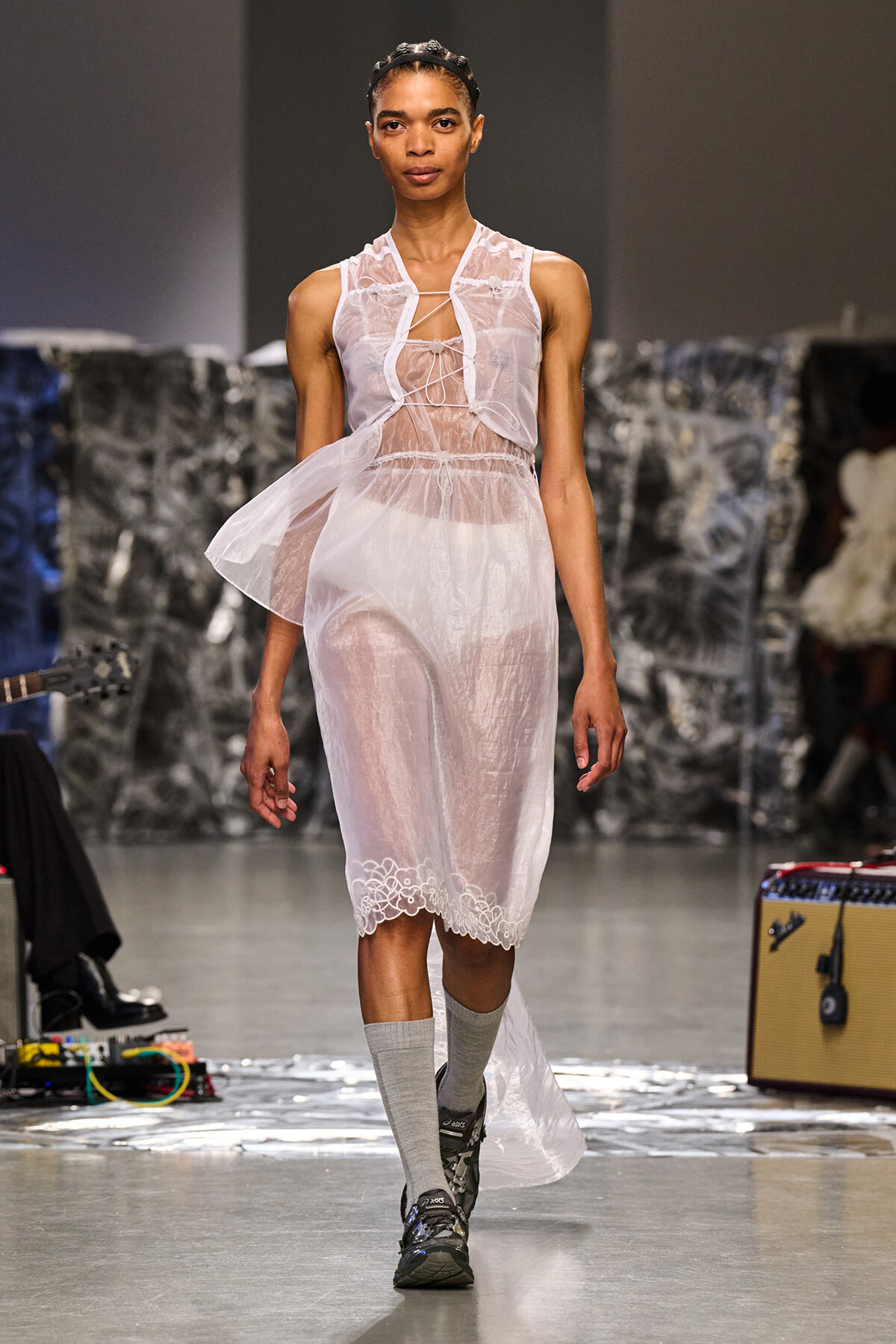 Model walks a runway in a sheer white lace midi dress with a crisscross front tie, paired with gray socks and sneakers.