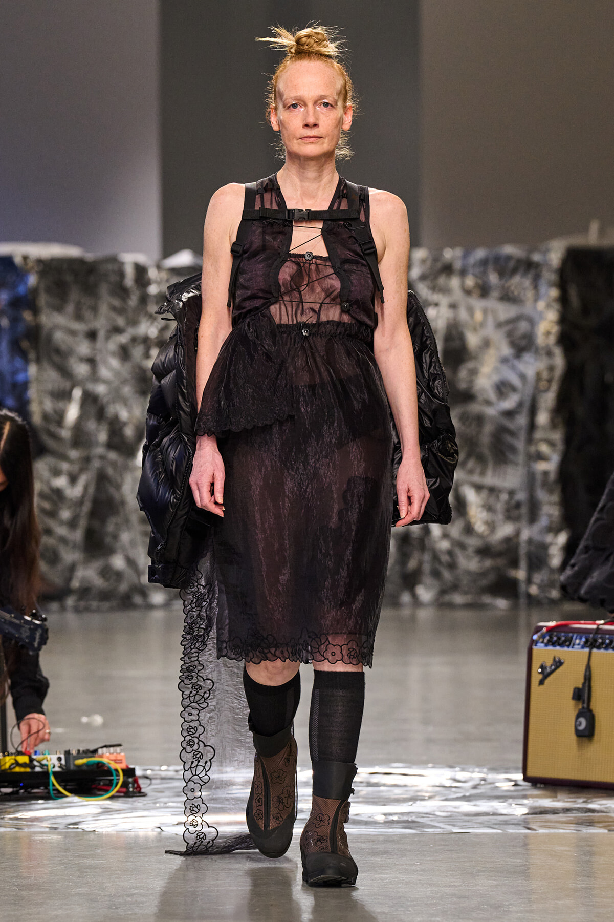 Model walking a runway in a sheer black lace midi dress with harness details and a black puffer jacket draped over her shoulders.