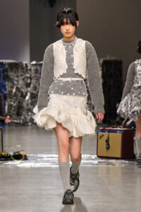 Model on a runway wearing a gray knit sweater with a white lace vest and a tiered, ruffled white skirt, headband, and gray socks with black shoes.