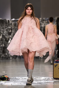 Model walking the runway in a pale pink floral lace, voluminous dress at a fashion show.