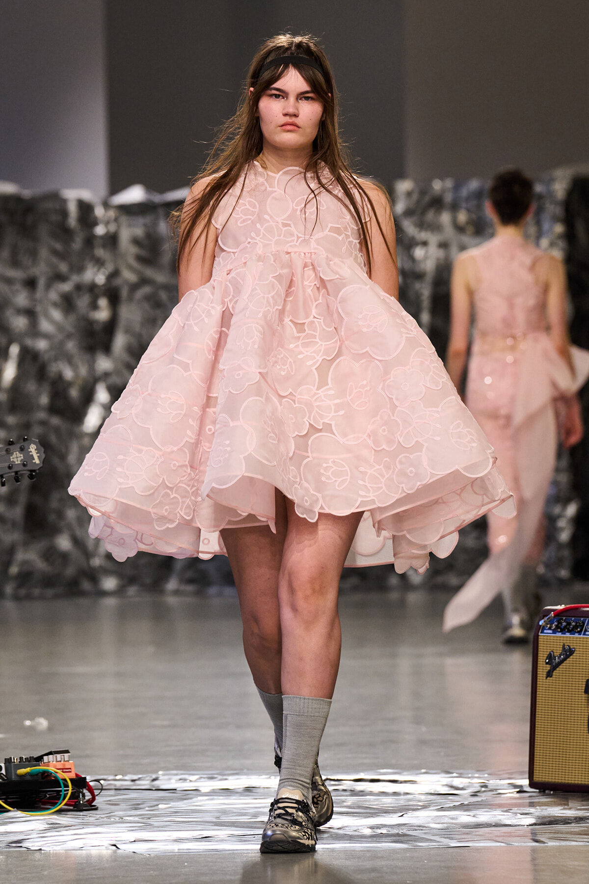 Model walking the runway in a pale pink floral lace, voluminous dress at a fashion show.