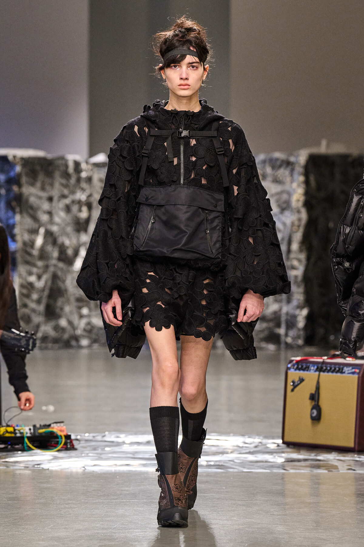 Model on a fashion runway wearing a black floral lace outfit with oversized sleeves, a harness, and a front black pouch. Boots and a headband complete the look.