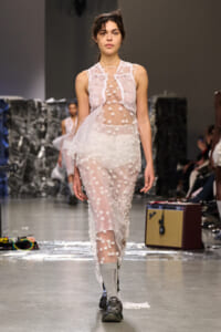 Model walking a fashion show in a translucent white dress with polka-dot floral appliqués and lace, paired with gray socks and sneakers.