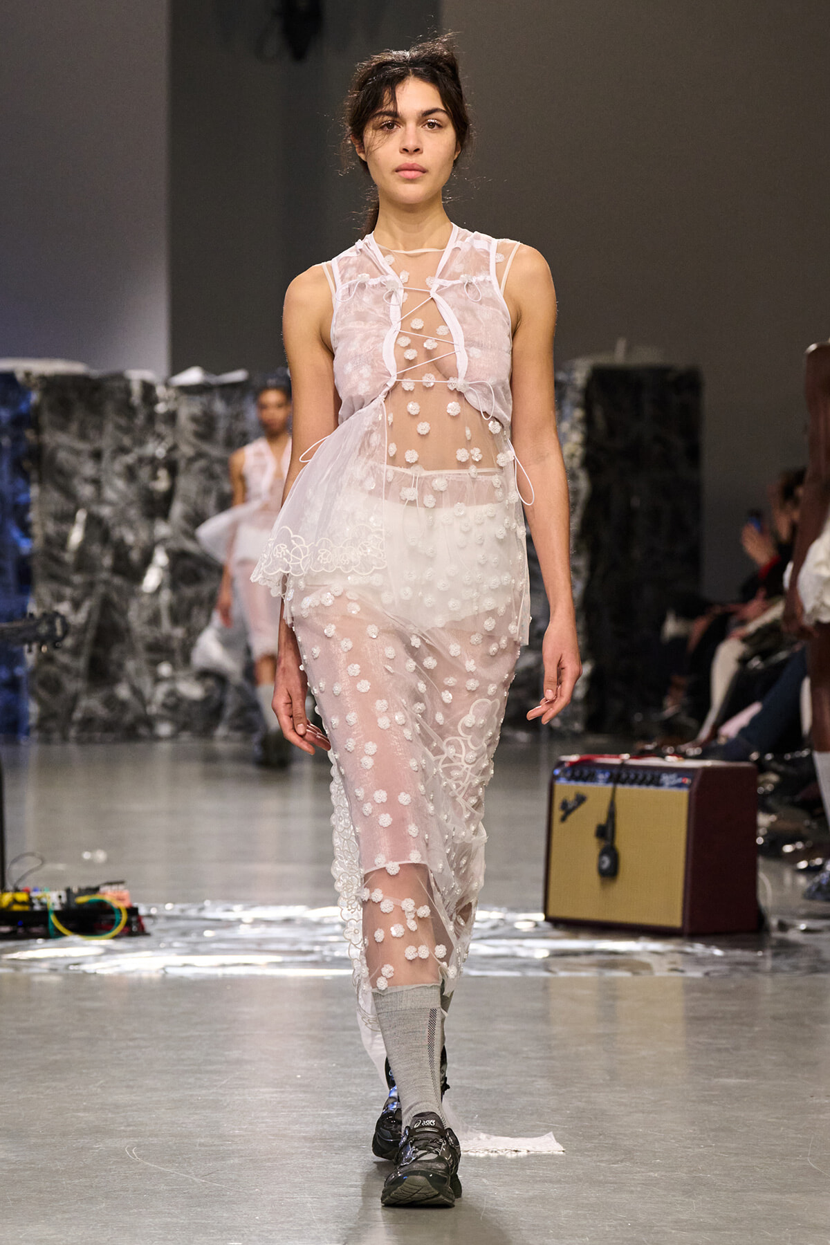 Model walking a fashion show in a translucent white dress with polka-dot floral appliqués and lace, paired with gray socks and sneakers.
