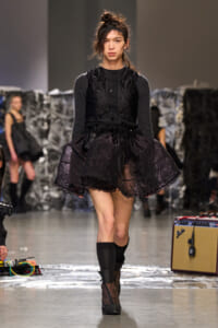 Model on a runway wearing a black lace layered dress over a dark knit top, with a voluminous skirt and black boots.
