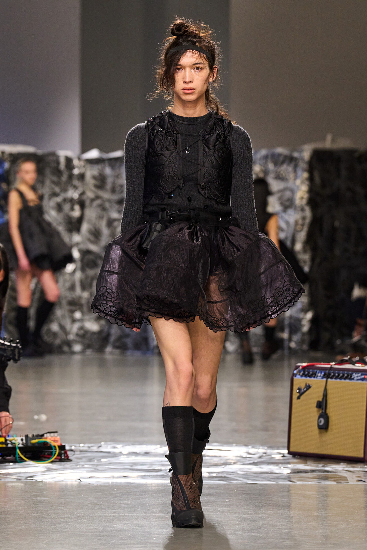 Model on a runway wearing a black lace layered dress over a dark knit top, with a voluminous skirt and black boots.