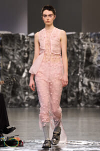 Runway model in a pale pink, sheer dress with 3D pink floral appliques, walking forward on a fashion show runway with gray backdrop.