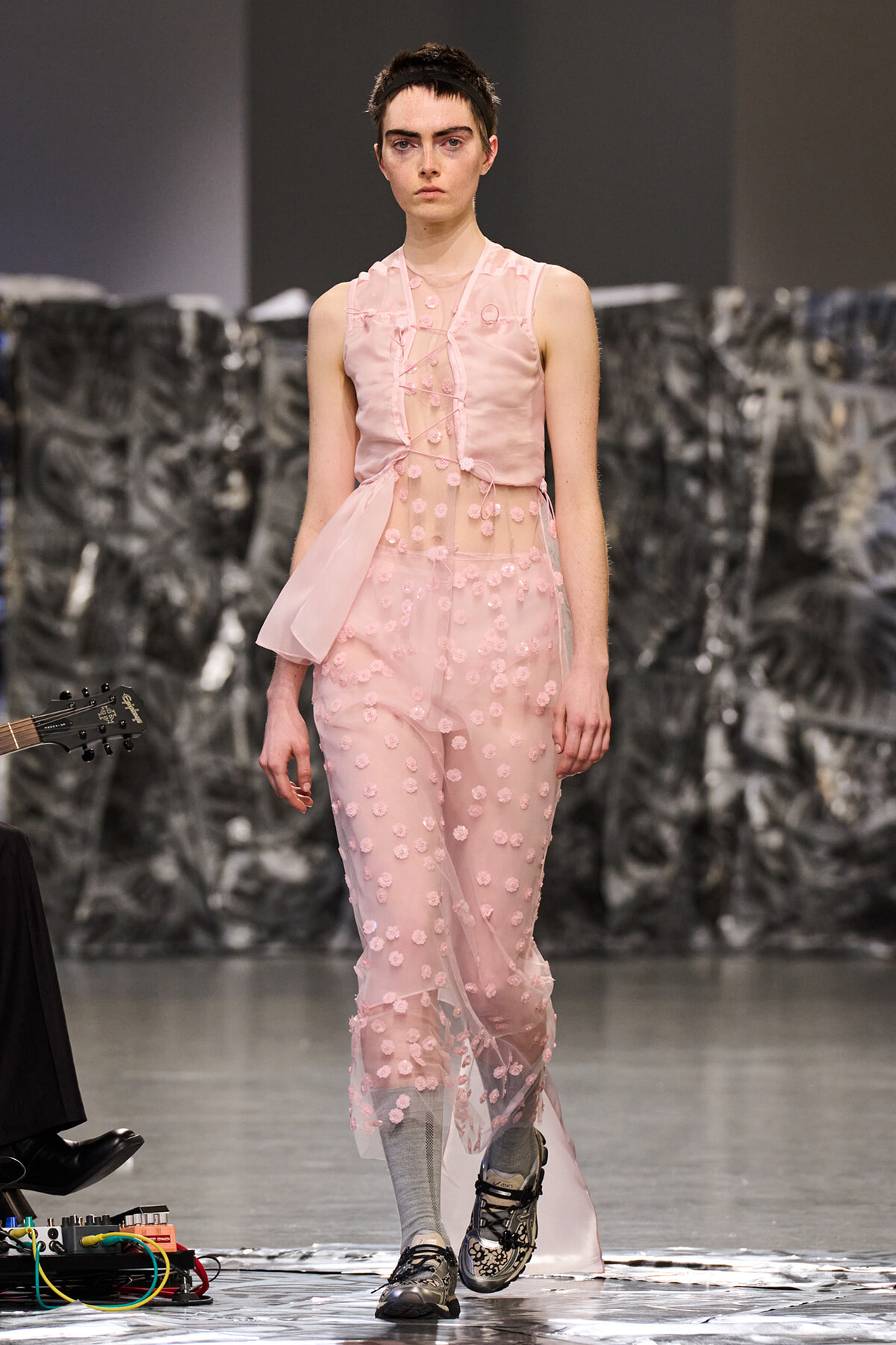 Runway model in a pale pink, sheer dress with 3D pink floral appliques, walking forward on a fashion show runway with gray backdrop.