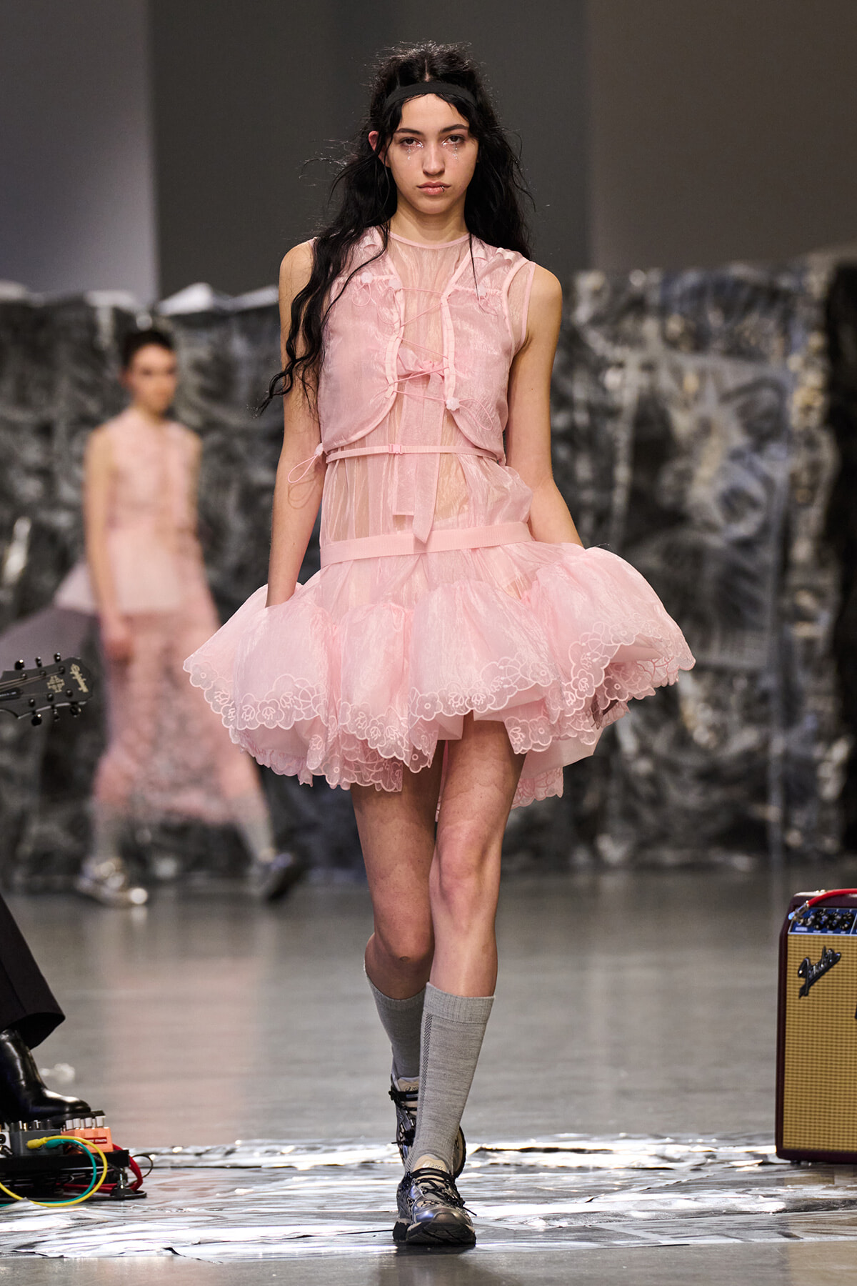 Model walking the runway in a pale pink lace tutu dress with a sheer bodice, ribbon accents, and a matching belt, paired with gray knee-high socks and dark shoes.