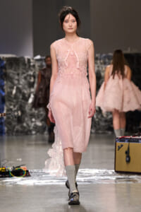 Model walks a fashion show in a pale pink lace dress with floral appliqué and sheer overlay on a runway.