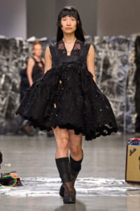 Model walks runway in a voluminous black lace dress with floral appliqués and a harness, headband, and sheer top.
