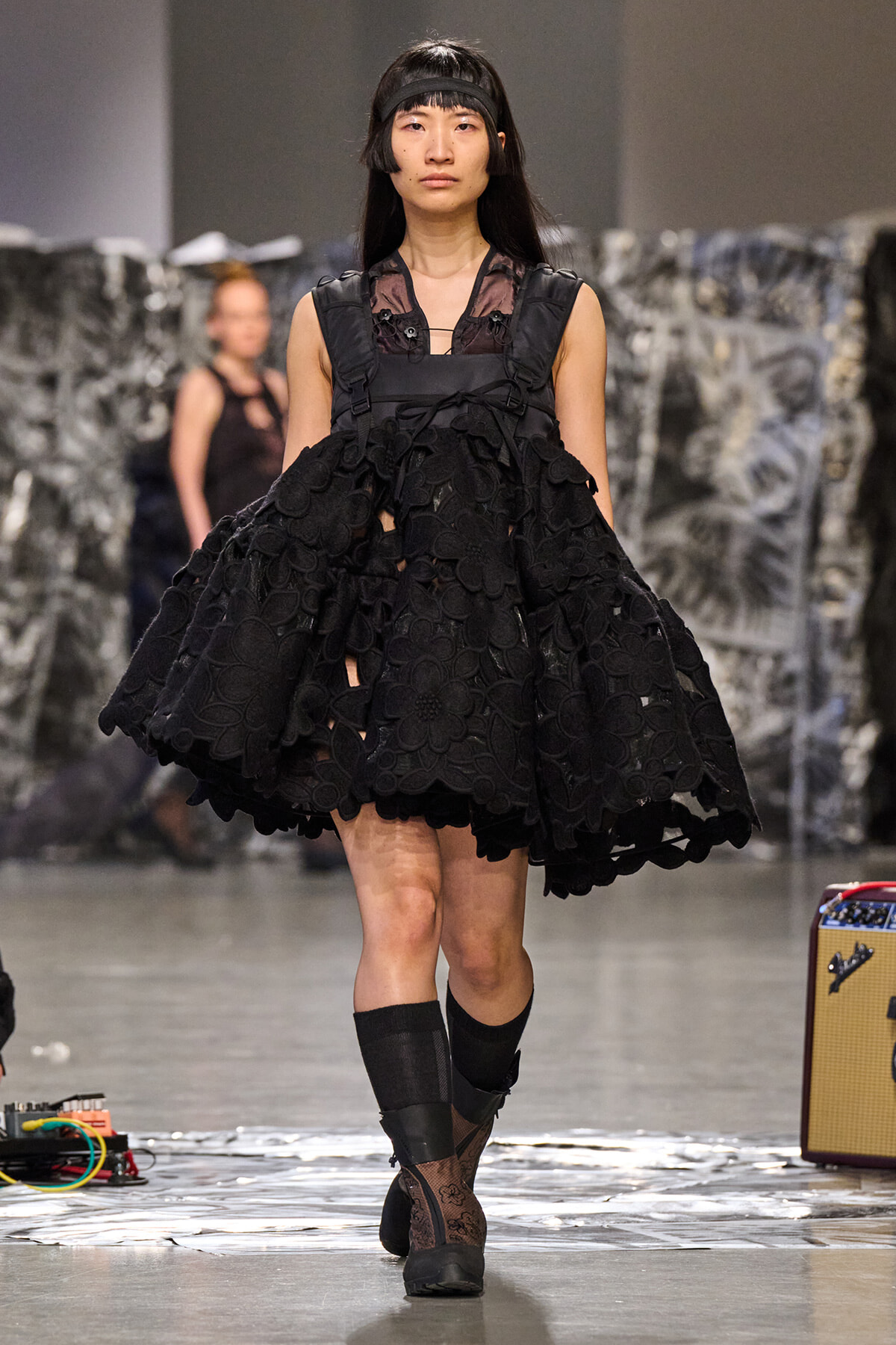Model walks runway in a voluminous black lace dress with floral appliqués and a harness, headband, and sheer top.
