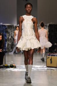 Model walks a runway in a cream quilted sleeveless dress with a ruffled, scalloped hem and wide belt details.