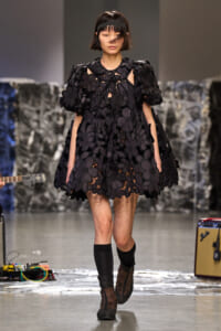 Model walking runway in a black voluminous floral dress with oversized 3D flower appliqués and a headband.