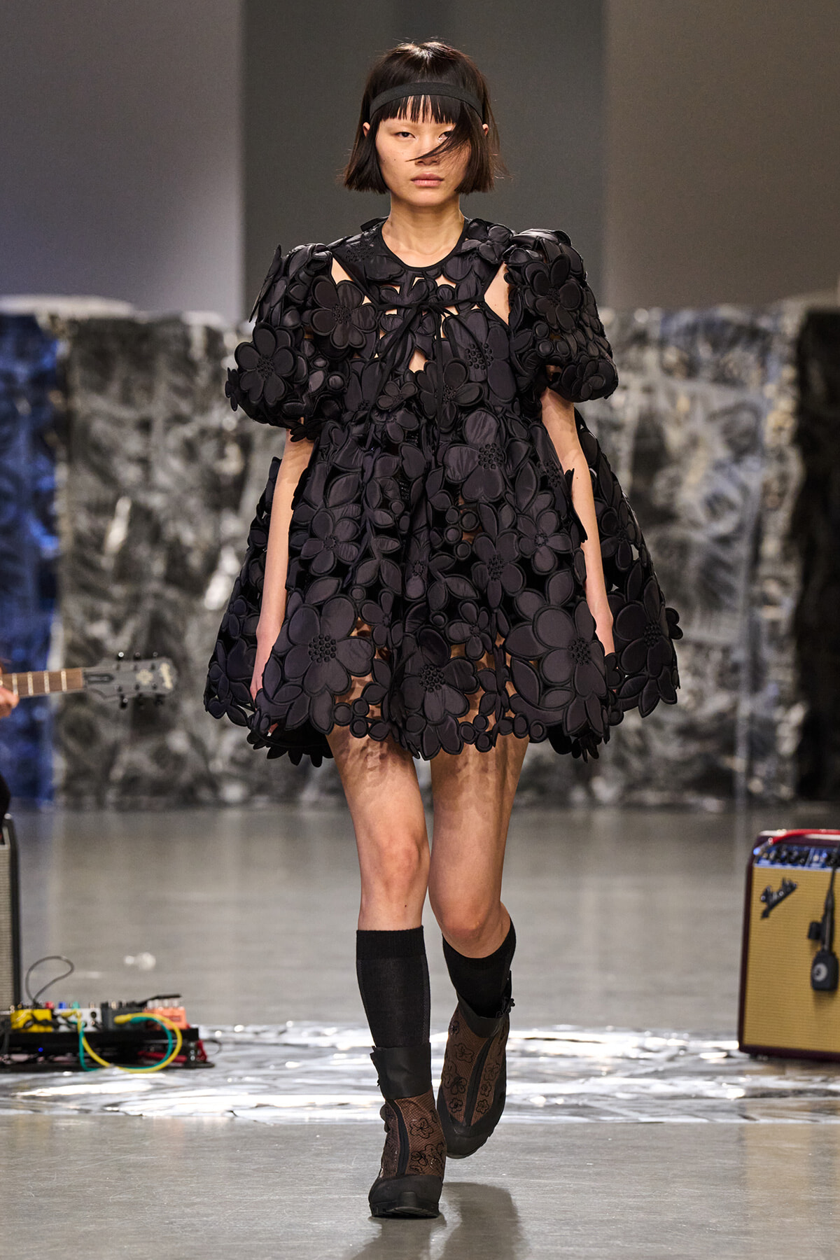 Model walking runway in a black voluminous floral dress with oversized 3D flower appliqués and a headband.