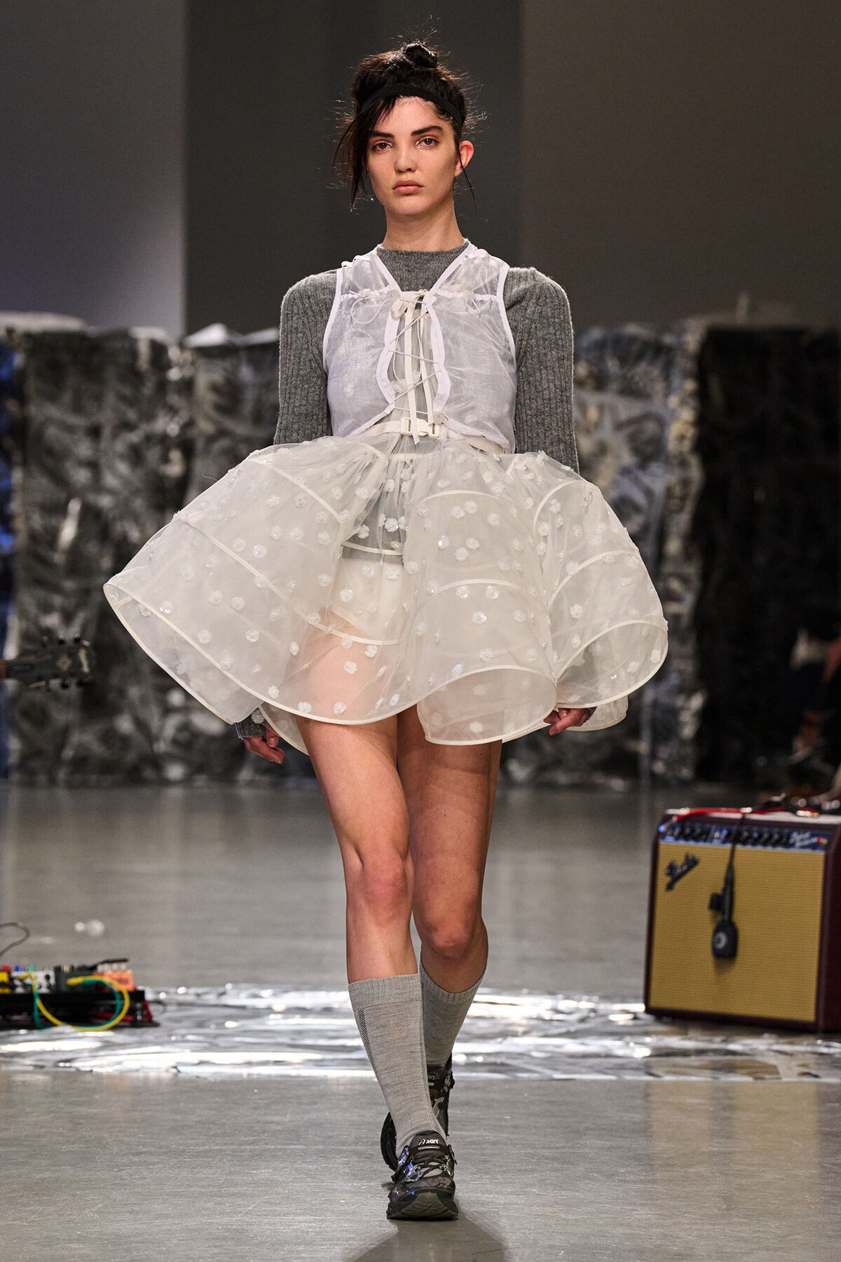 Model on a runway wearing a sheer white, polka-dot layered dress over a gray knit top, with gray knee-high socks and black sneakers, headband included.